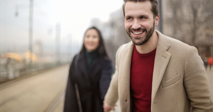 Smiling man in a camel coat and dark red sweater walking hand in hand with a woman along a hazy winter street