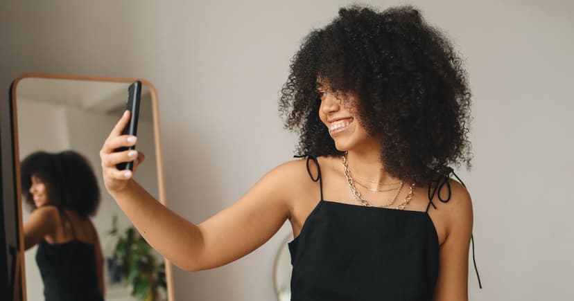 Smiling woman in a black dress taking a photo of herself with her phone in front of a full-length mirror at home