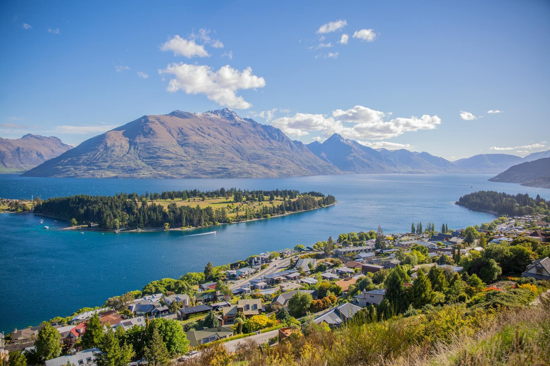 A lakeside town below brown mountains, with a wooded peninsula under a blue sky.