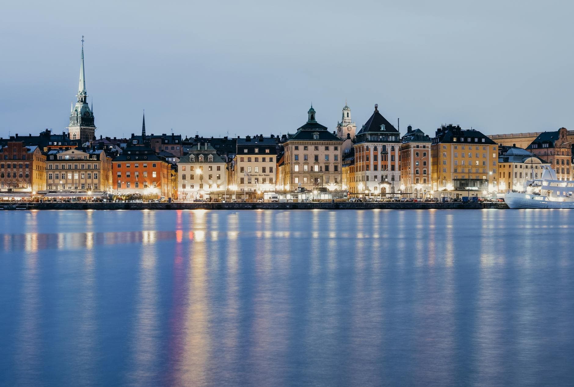 Upplysta fasader vid vattnet i skymning speglas i den lugna ytan intill en vit båt.