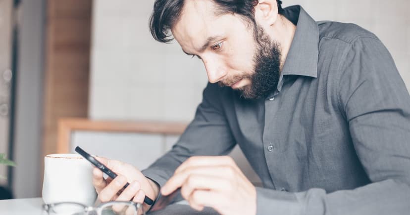 Bearded man in a grey shirt frowning at his smartphone over a coffee at a table