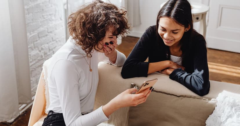 Woman showing a message on her phone to a friend leaning over the back of a sofa in a bright living room