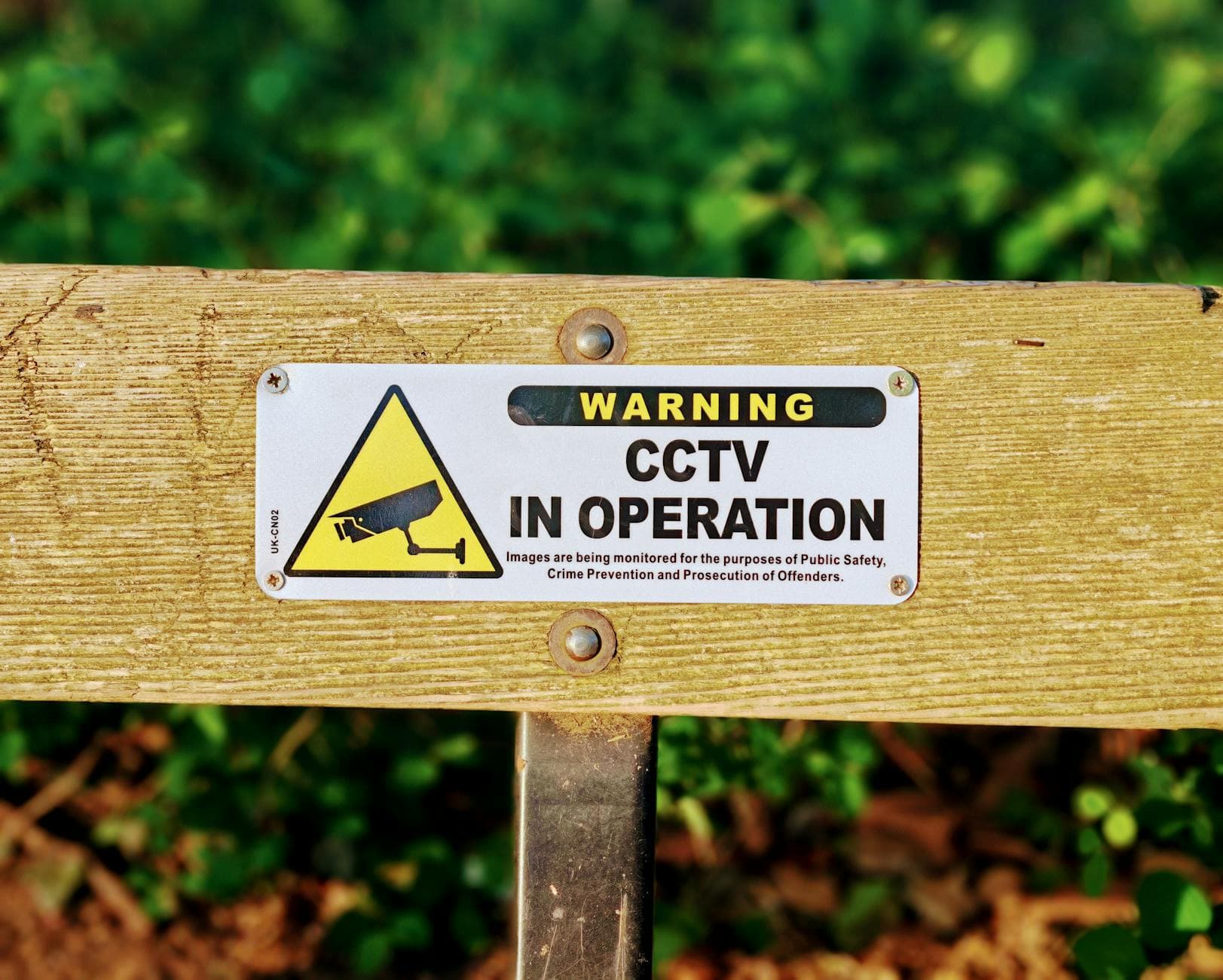 A yellow and white CCTV warning sign screwed to a wooden rail in front of green leaves.