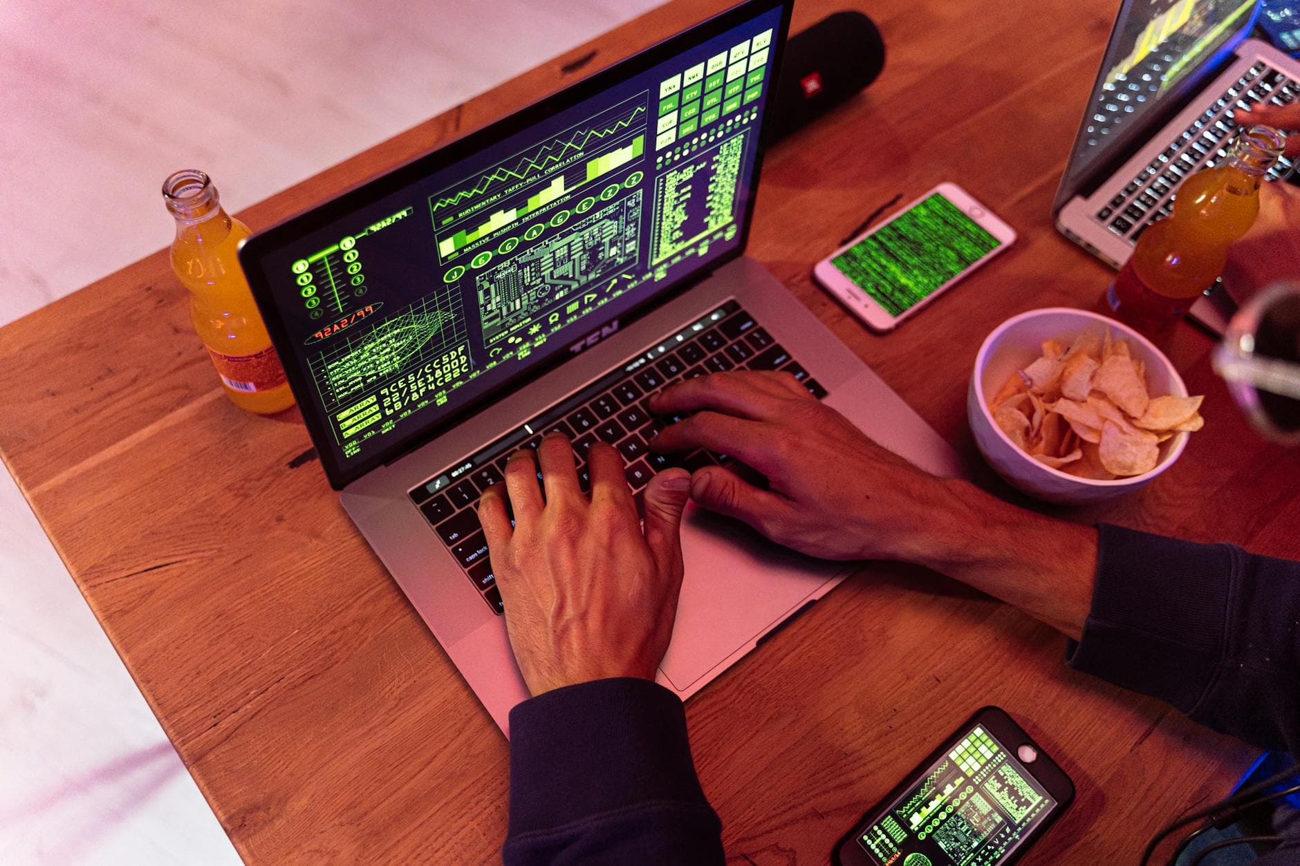 Hände tippen auf einem Laptop mit grünem Code, daneben Handy, Chips und Limoflasche.