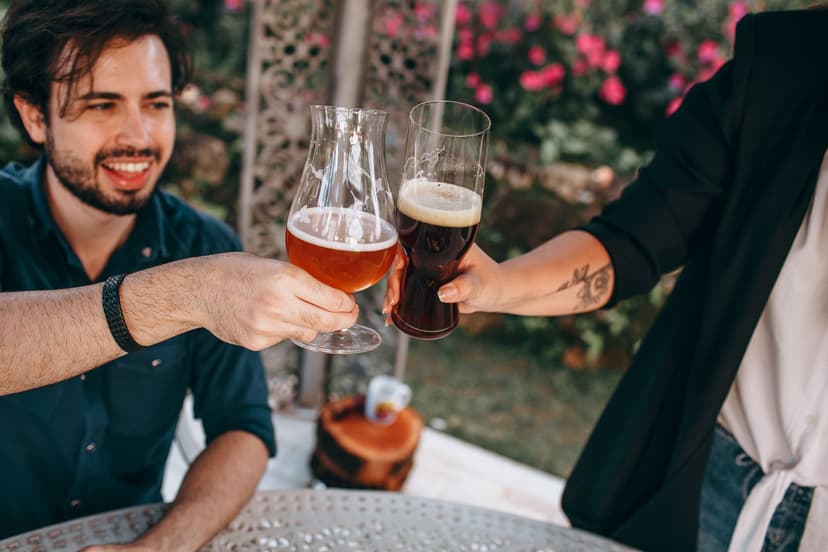 Lächelnder Mann mit Bart und eine Frau stoßen an einem sommerlichen Gartentisch mit zwei Craft-Bier-Gläsern an, im Hintergrund blühen pinke Blumen