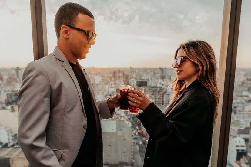 Elegantes Paar in Blazern stößt bei Sonnenuntergang in einer Rooftop-Bar mit Drinks an, hinter den Panoramafenstern erstreckt sich die Skyline der Stadt