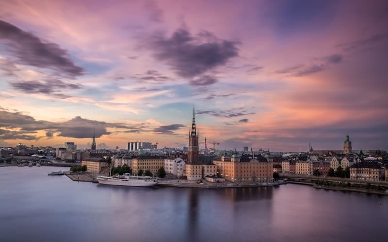 Stadssiluett med kyrkspira vid vattnet under rosa och lila kvällsmoln.