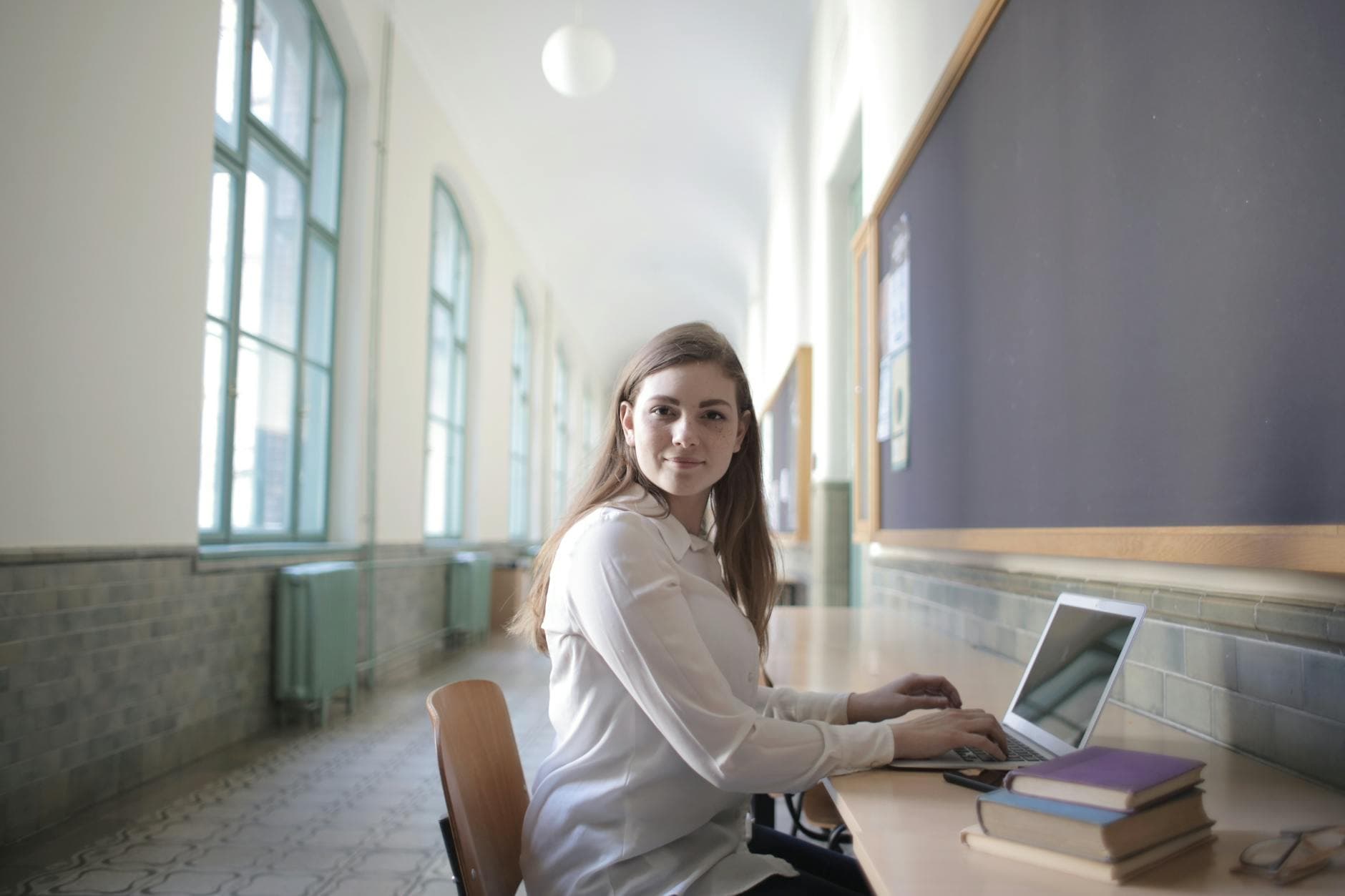 Kvinne med langt hår i hvit bluse sitter med laptop i en lys skolekorridor.
