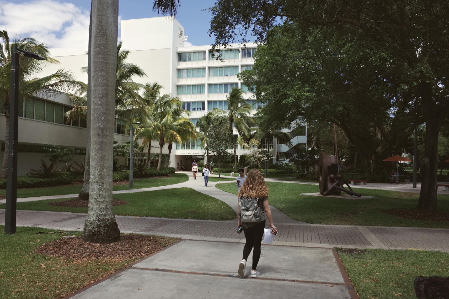 A person with a backpack walks a campus path between palms toward a white building.