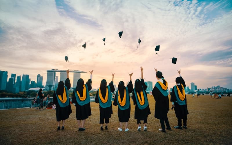 Zeven afgestudeerden in zwarte toga's gooien hun baretten omhoog op een grasveld.