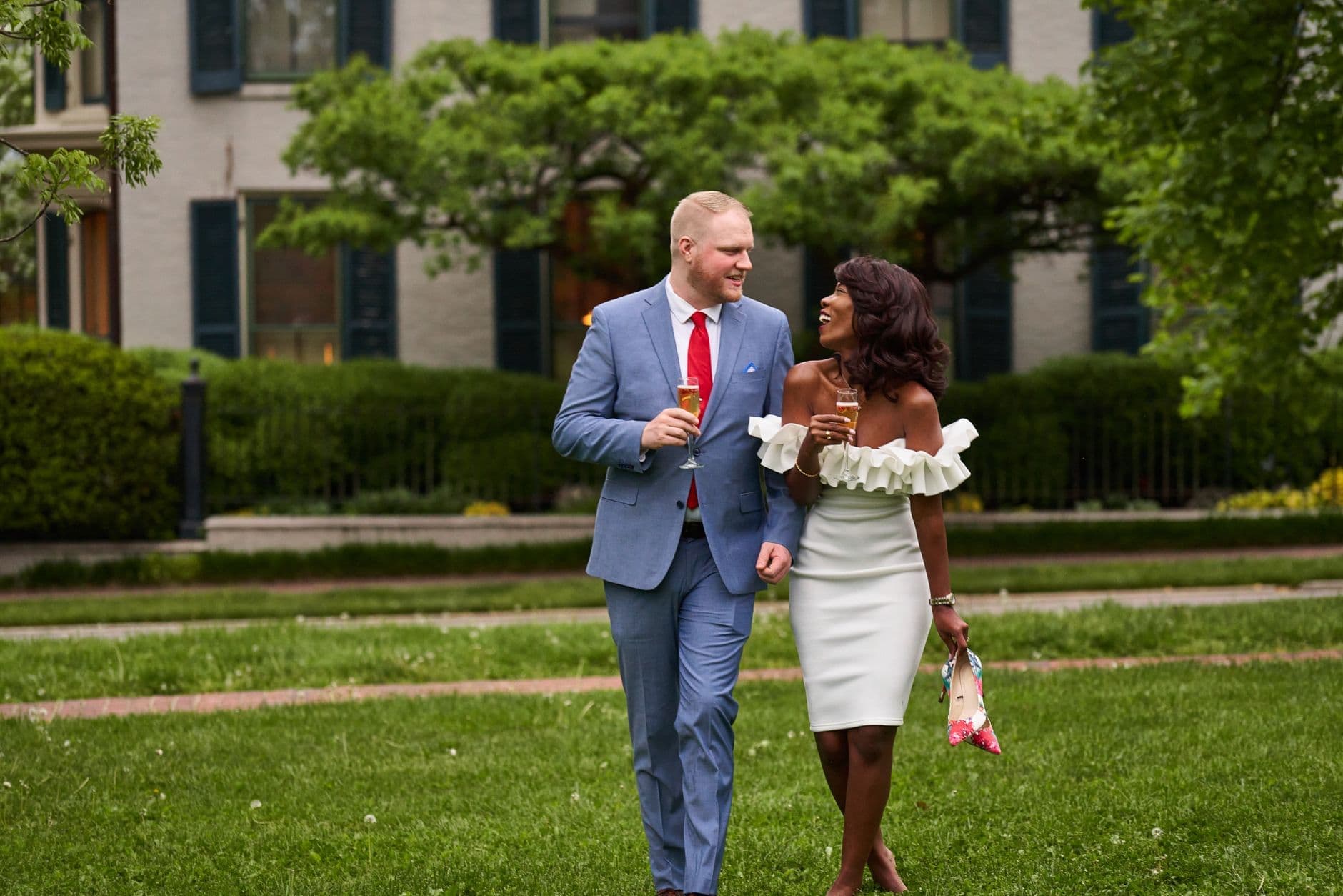 A man in a light blue suit and a woman in a white dress carry drinks across a lawn.