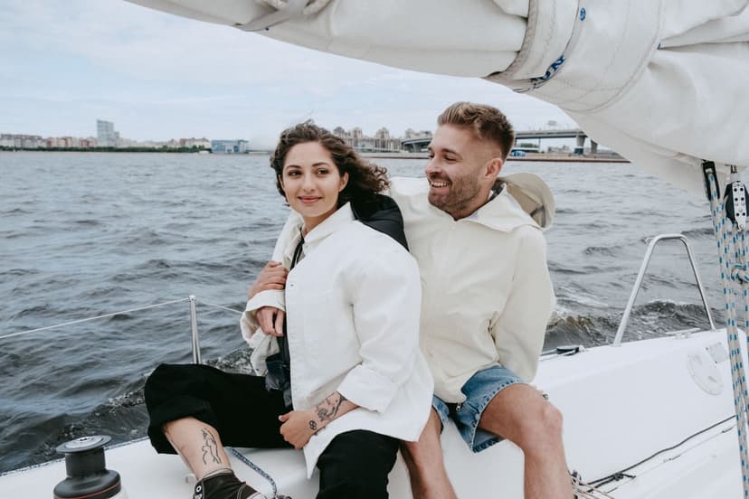 Smiling couple in light summer jackets sitting close together on the deck of a sailboat under a furled sail, with open water and a city skyline behind them