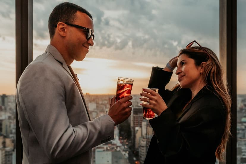 Elegant couple in blazers sharing iced cocktails by the floor-to-ceiling windows of a rooftop bar, with a golden sunset glowing over the city skyline below