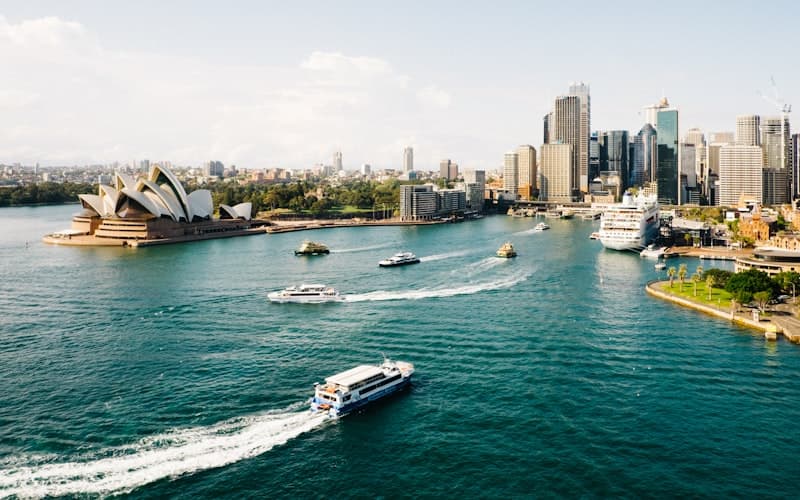 Ferries cross a blue harbour past a sail roofed opera house and a skyline of towers.