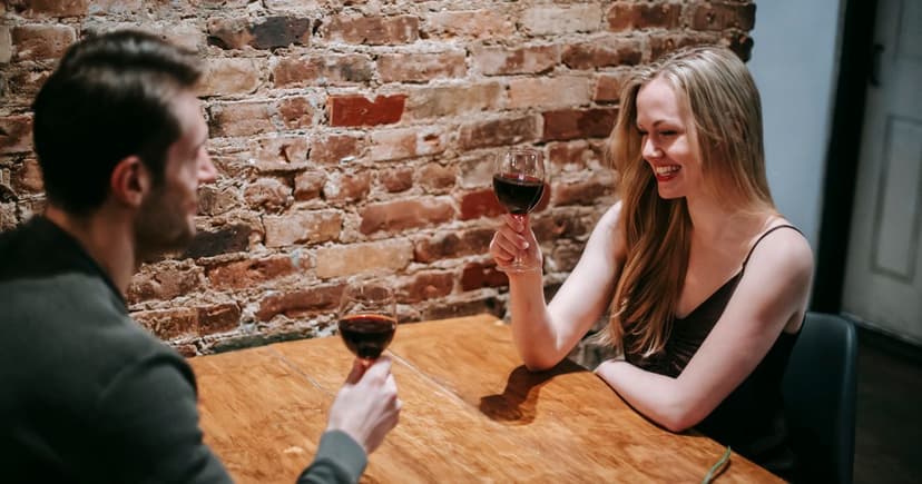 A man in a dark sweater and a blonde woman raise red wine glasses across a wooden table by exposed brick.