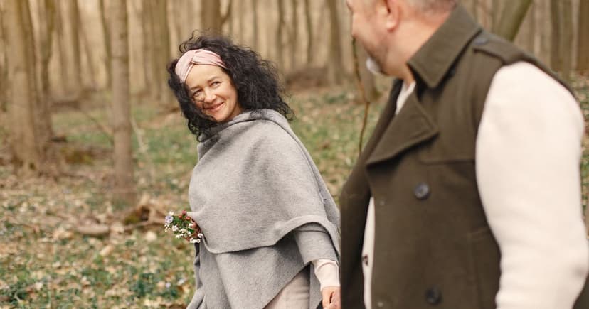 A smiling woman in a gray wool wrap looks back at a man in a green coat while walking through bare woods.