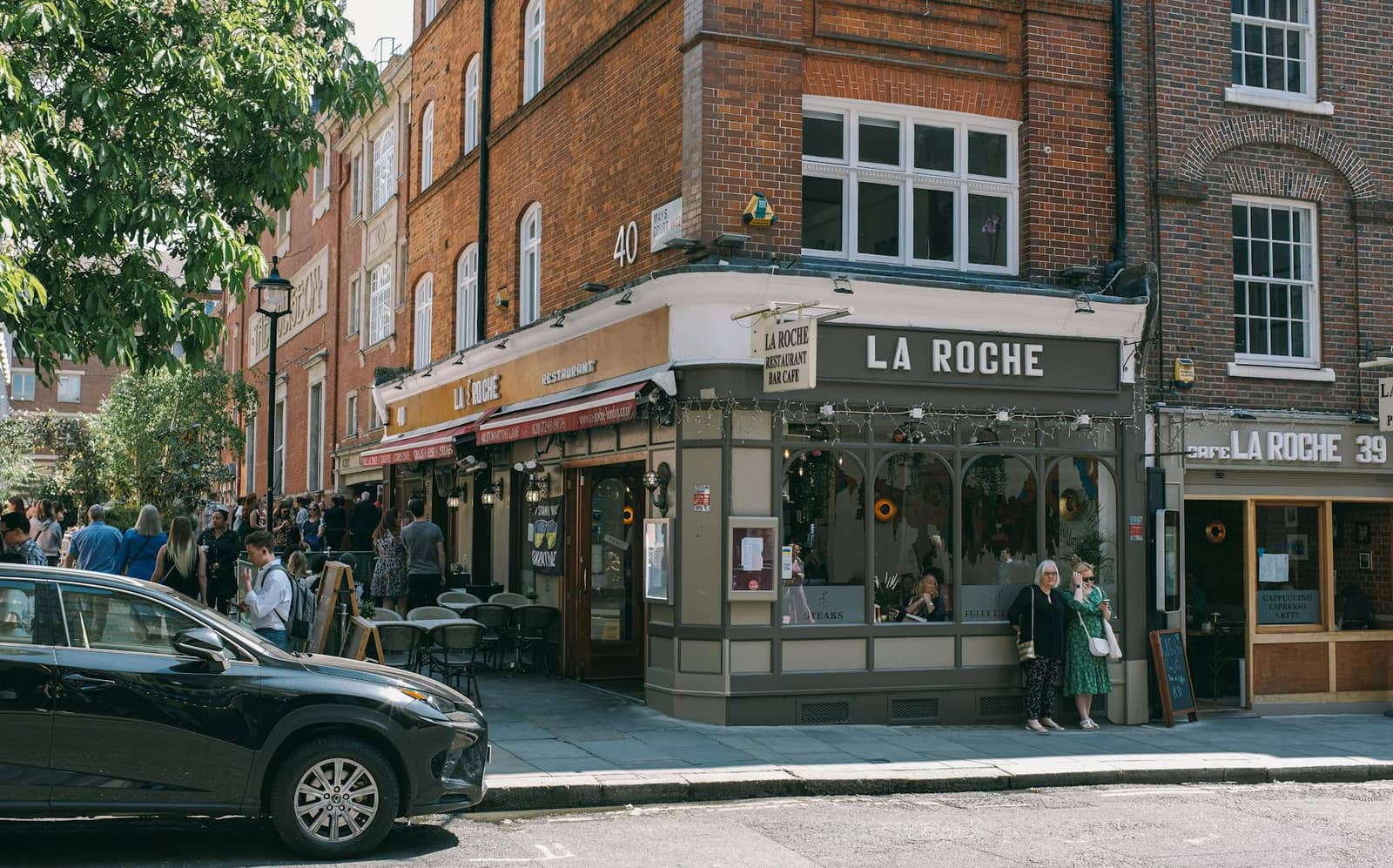 People stand outside a red brick corner cafe on a sunny street with a car parked nearby.