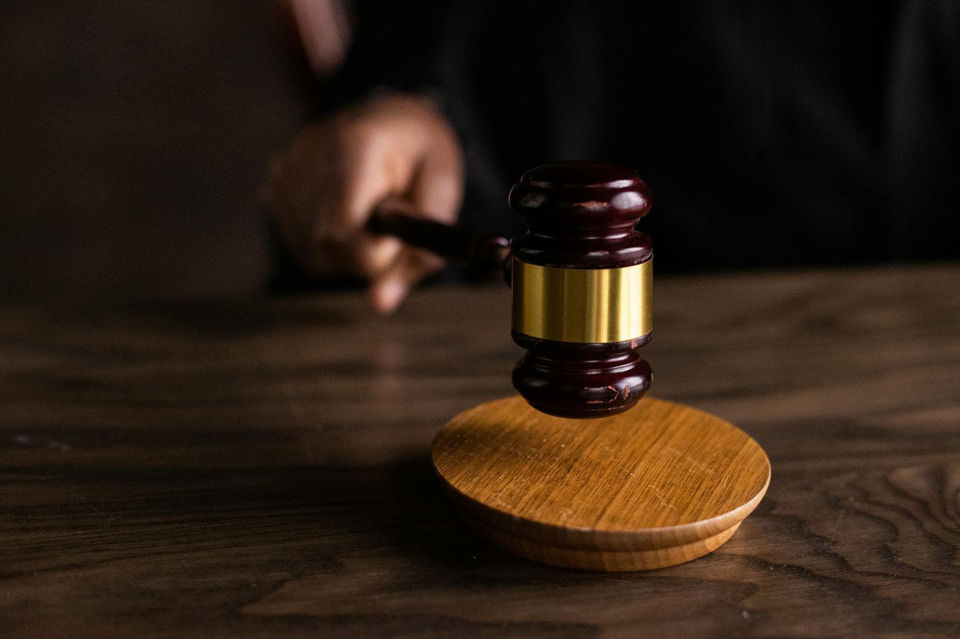 A wooden gavel rests on its block on a dark desk with a hand blurred behind.