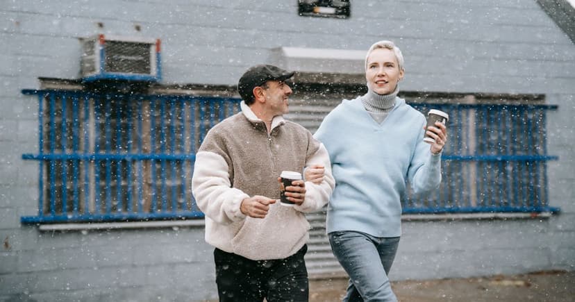 A man in a fleece pullover and a woman in a pale blue sweater walk with takeout cups through falling snow.