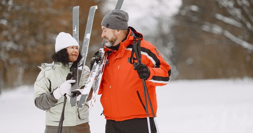 A woman in a white knit hat and a man in an orange jacket carry skis and poles across snowy ground.