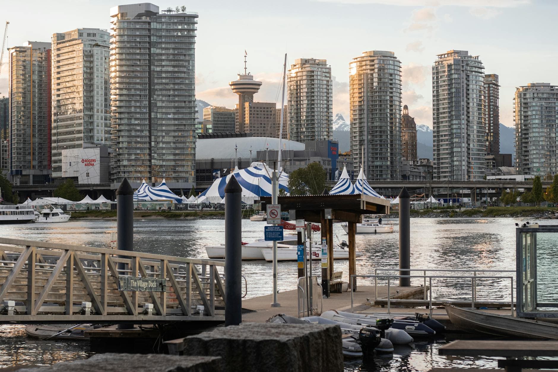 Glass towers rise behind a marina with sailboats and blue striped festival tents.