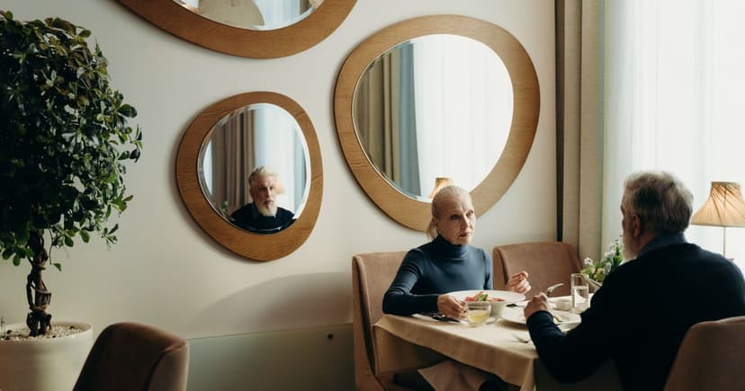 An older woman in a navy turtleneck dines opposite a grey-haired man in a bright restaurant with round wall mirrors.