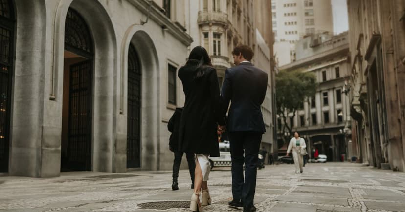 A couple in a navy suit and a black coat walk hand in hand away from the camera down a stone-paved street lined with tall, columned classical buildings.