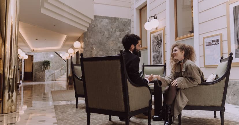 A well-dressed man and woman sit facing each other in armchairs in a marble hotel lobby, talking and smiling across a small table.