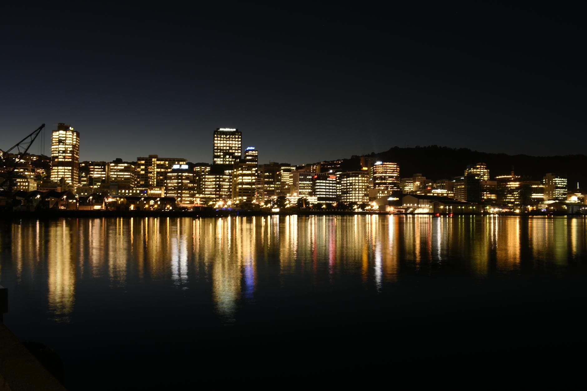Golden office lights along a waterfront reflect in calm harbour water after dark.