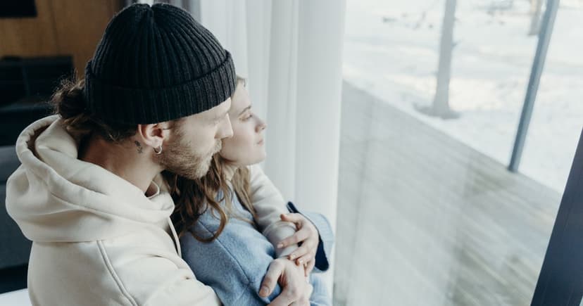 A man in a cream hoodie and a knitted beanie sits close to a woman in a pale blue sweater at a tall window, both looking out at deep snow.