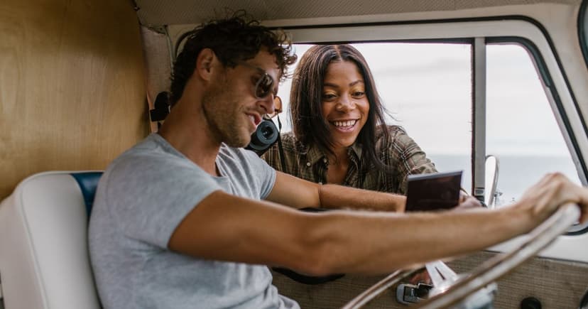 A man in a grey t-shirt sits at the wheel of an old van while a woman in a checked shirt leans in through the open window, both laughing over something they are reading.