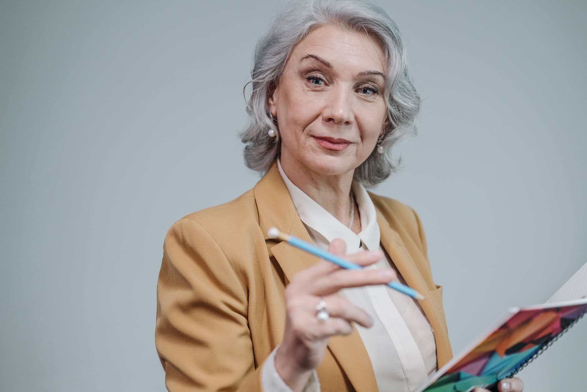 Žena s vlnitými šedivými vlasy v okrovém saku drží tužku a barevný blok.
