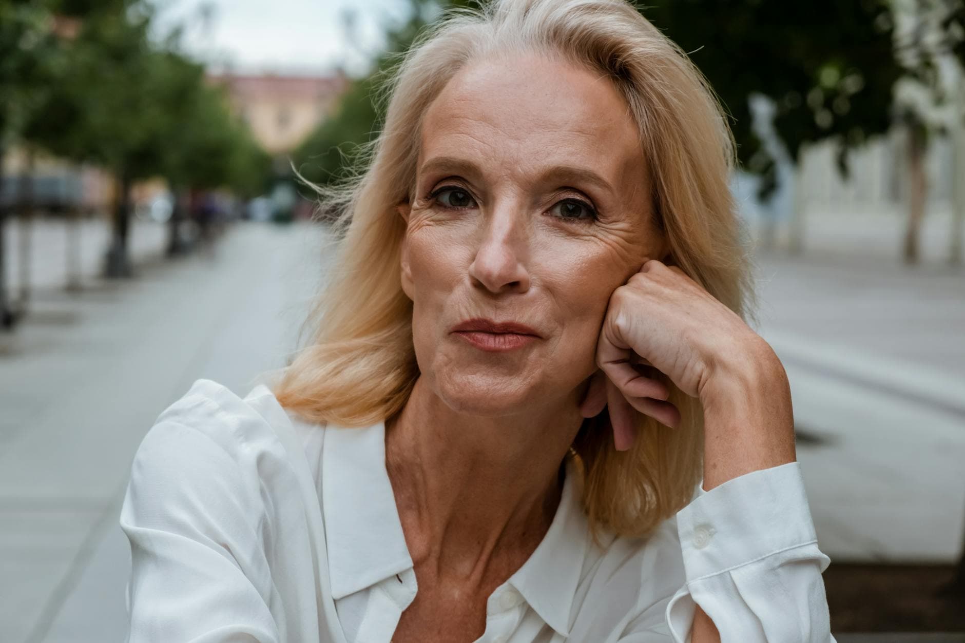 Blond kvinde i hvid skjorte hviler kinden i hånden på en træbeplantet gade.
