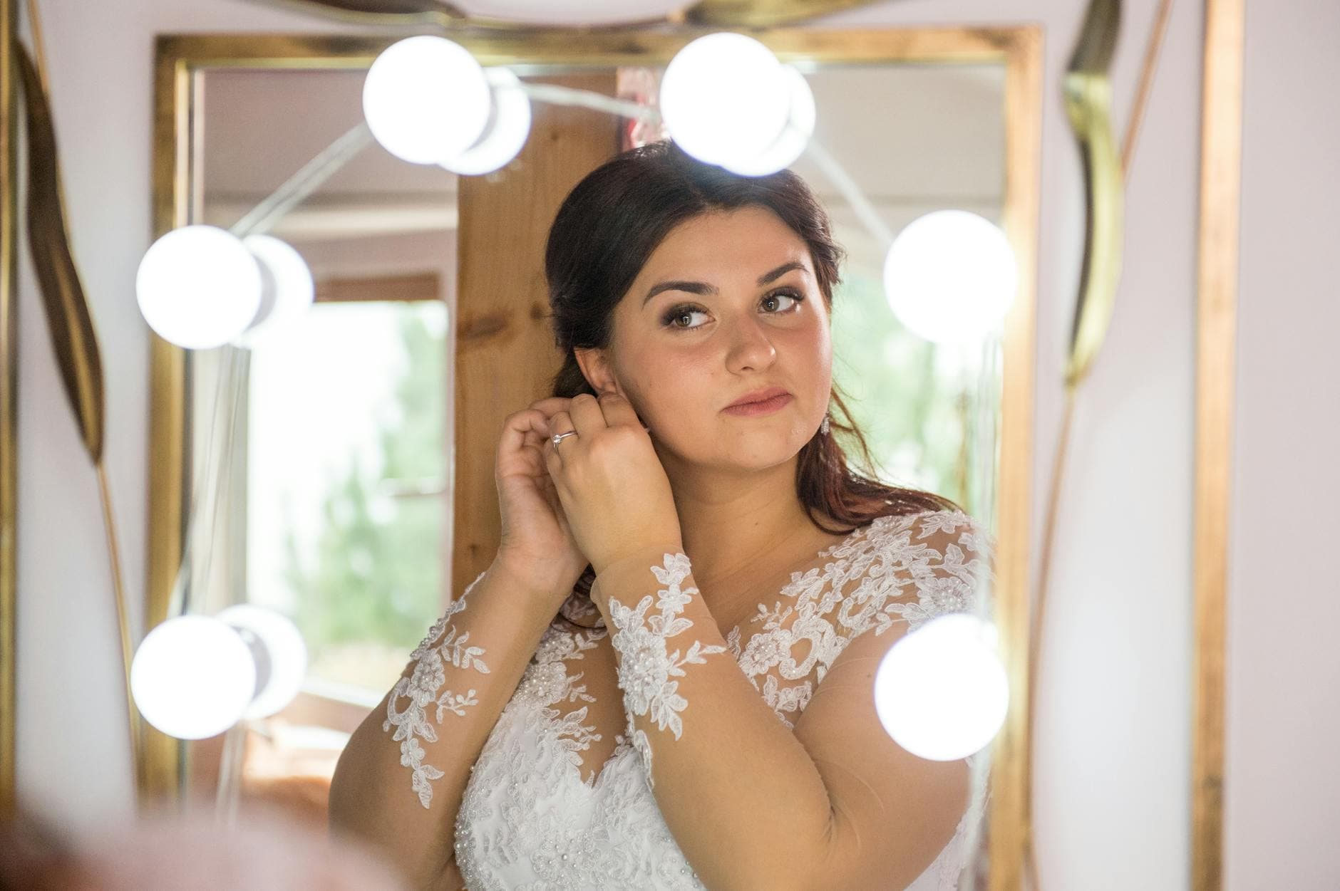 Vrouw in witte kanten jurk doet een oorbel in voor een spiegel met ronde lampen.