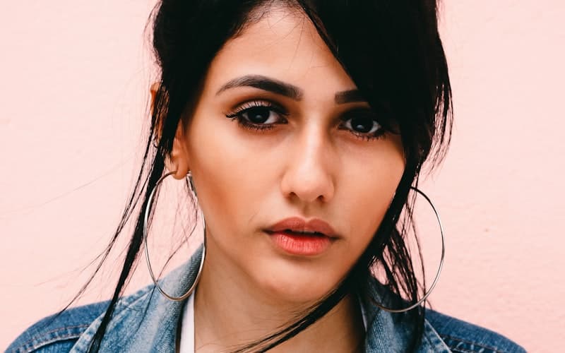 Nahaufnahme einer Person mit dunklen Haaren, großen Creolen und Jeansjacke vor Rosa.