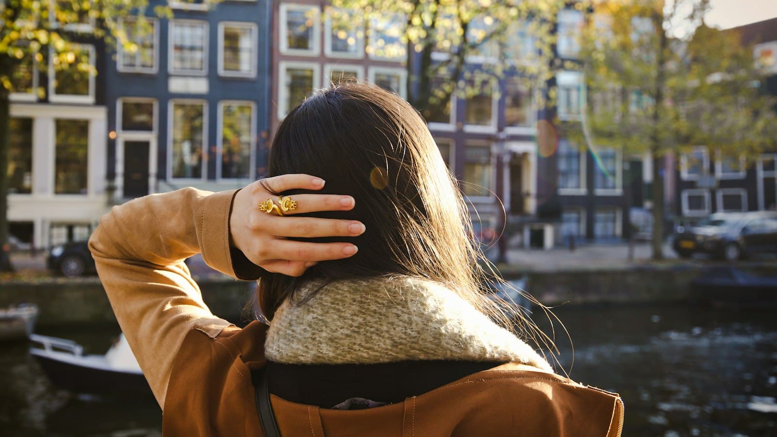Vrouw in een camelkleurige jas, van achteren gezien, kijkt in herfstlicht uit over een Amsterdamse gracht