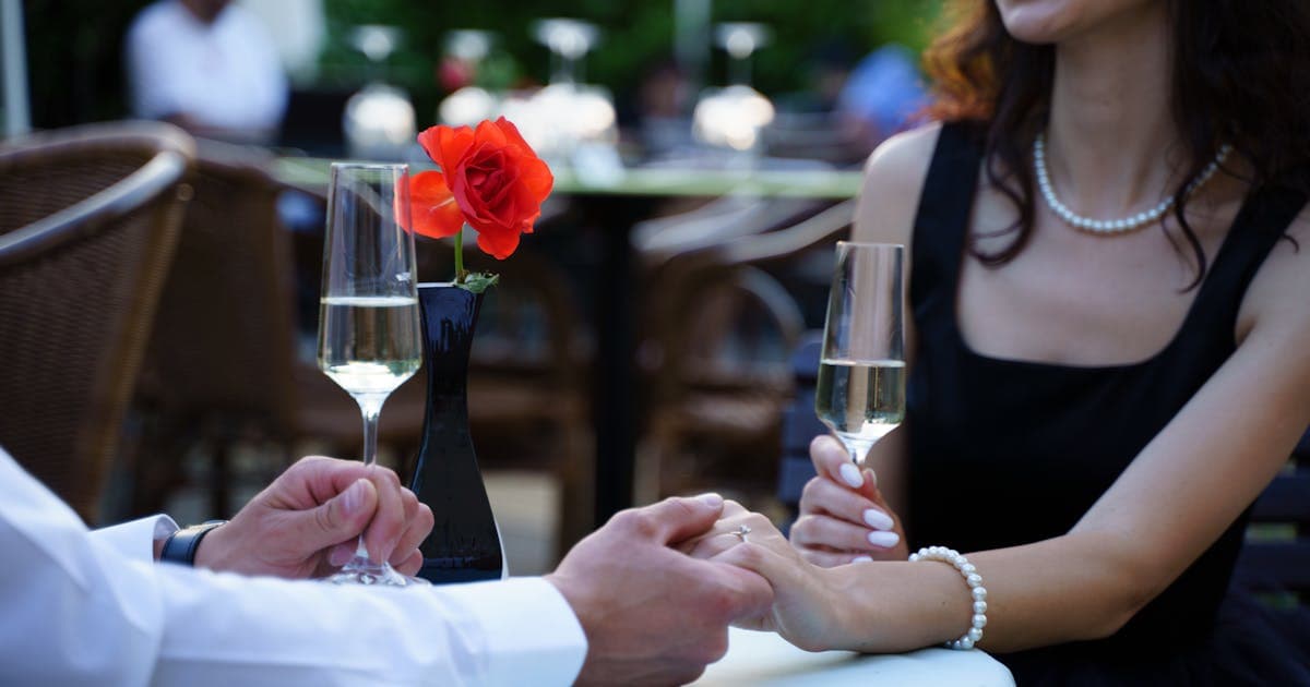To hænder mødes ved et bord med champagneglas og en rød rose i en mørk vase.