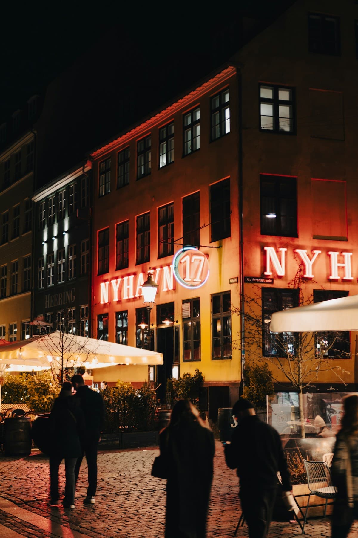 Rødt neonskilt på en gul facade lyser over brosten, mens folk går forbi om aftenen.