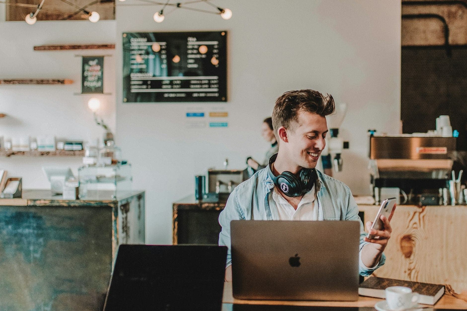 Lachende man met een koptelefoon om zijn nek kijkt op zijn telefoon achter een laptop in een koffiebar