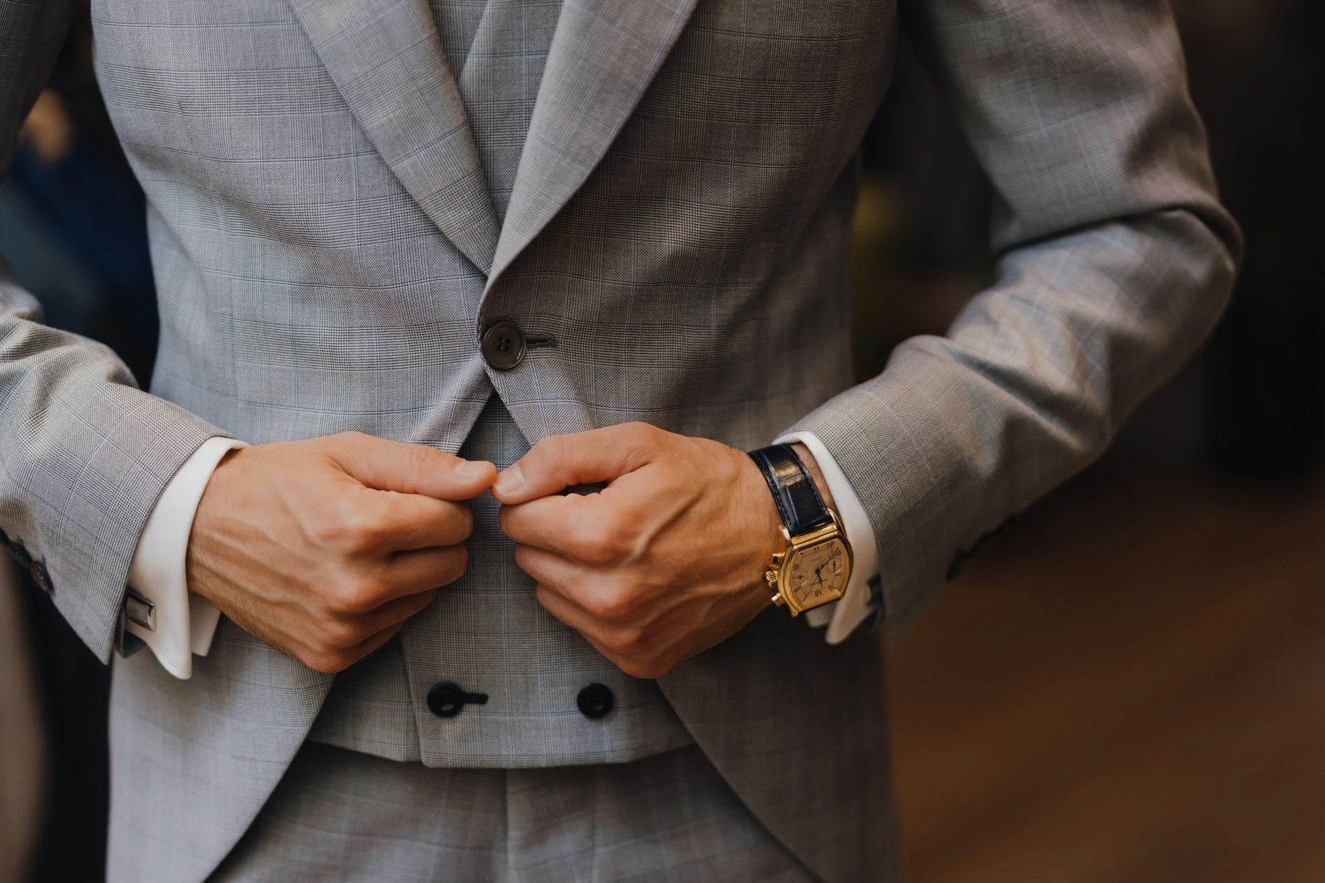 Man in a grey checked suit buttoning his jacket, with a gold watch on his wrist and his face out of frame