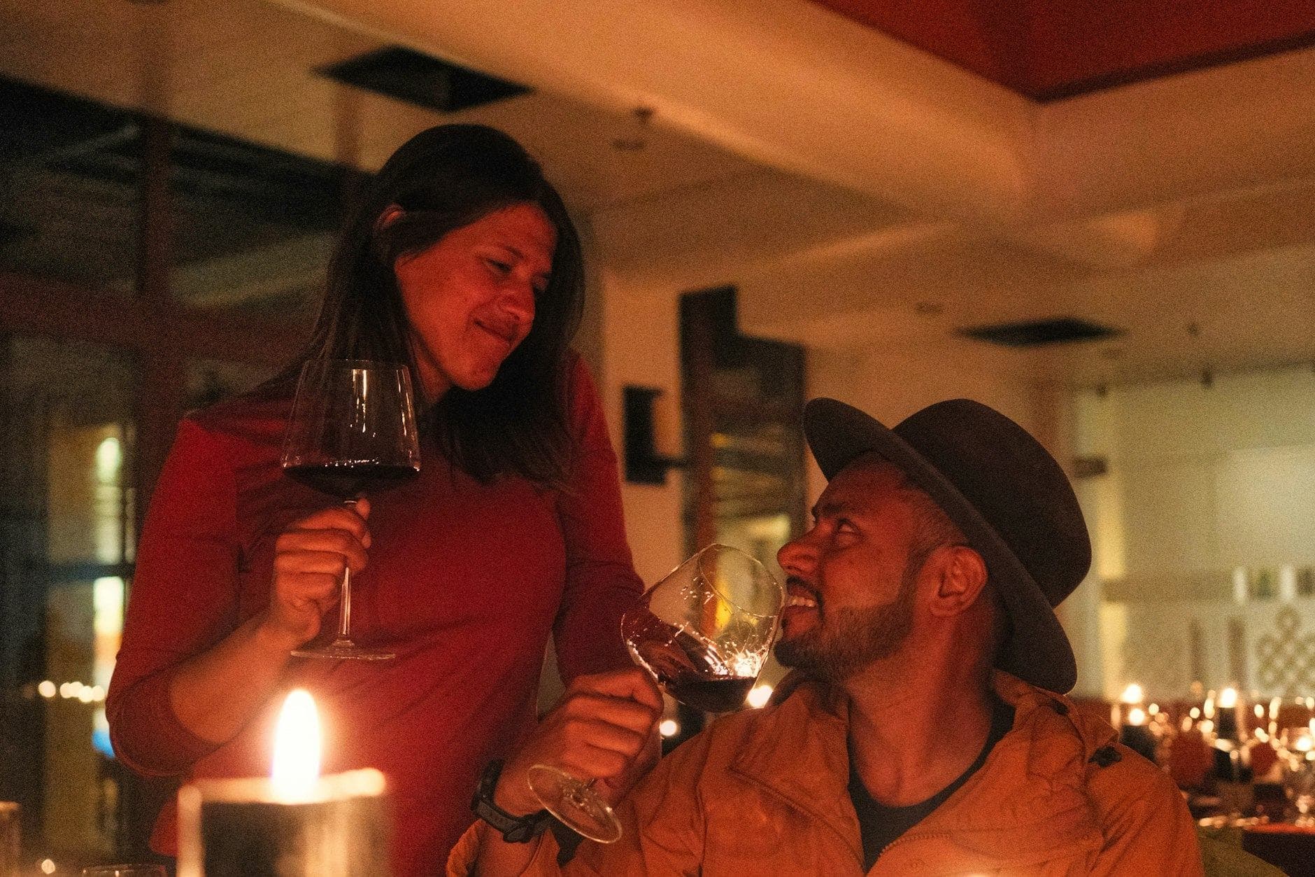 Lächelnde Frau in rotem Oberteil und Mann mit Hut stoßen bei Kerzenlicht im Restaurant mit Rotwein an
