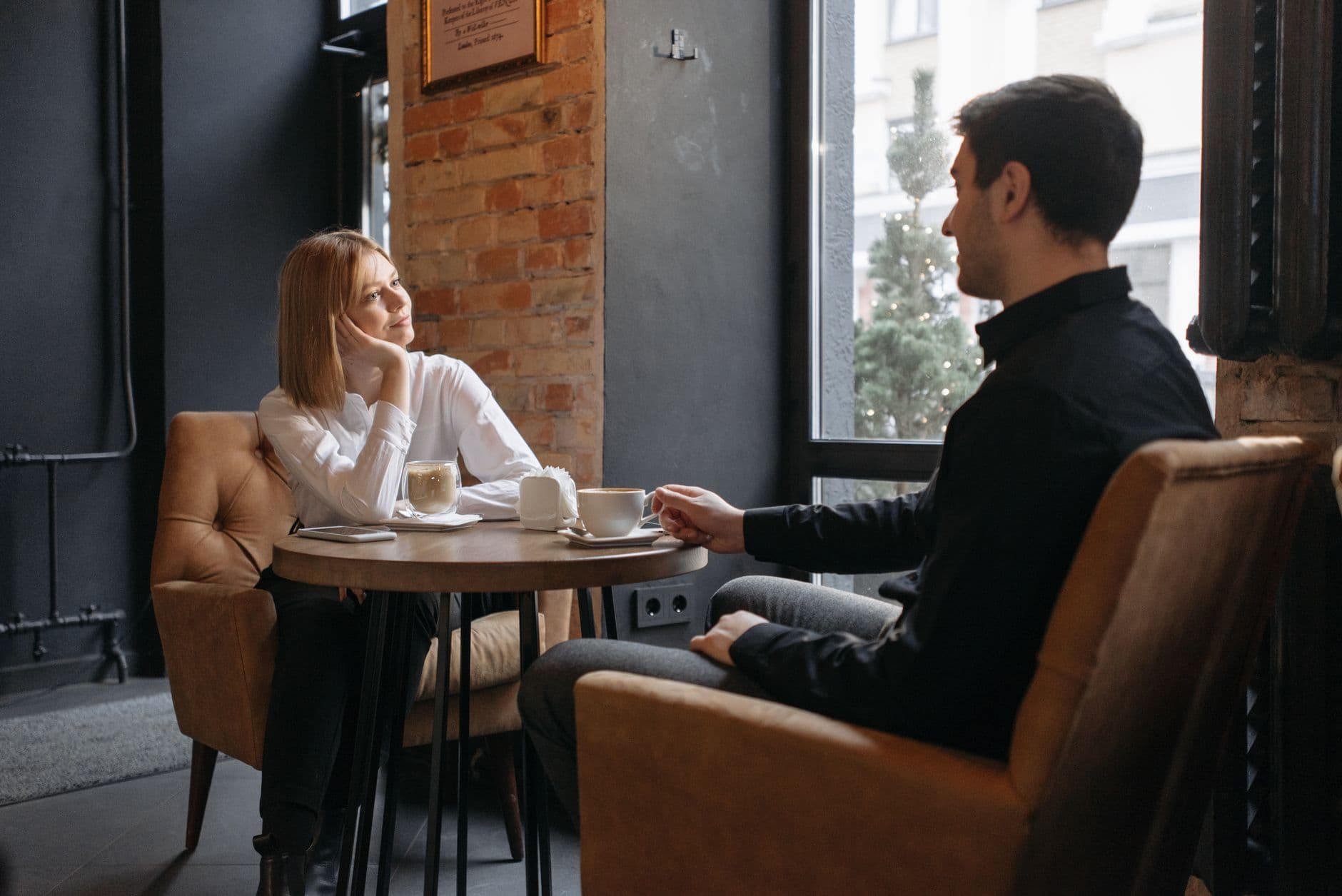 Lyshåret kvinde i hvid skjorte og mand i sort skjorte taler sammen over kaffe i en café med murstensvæg.