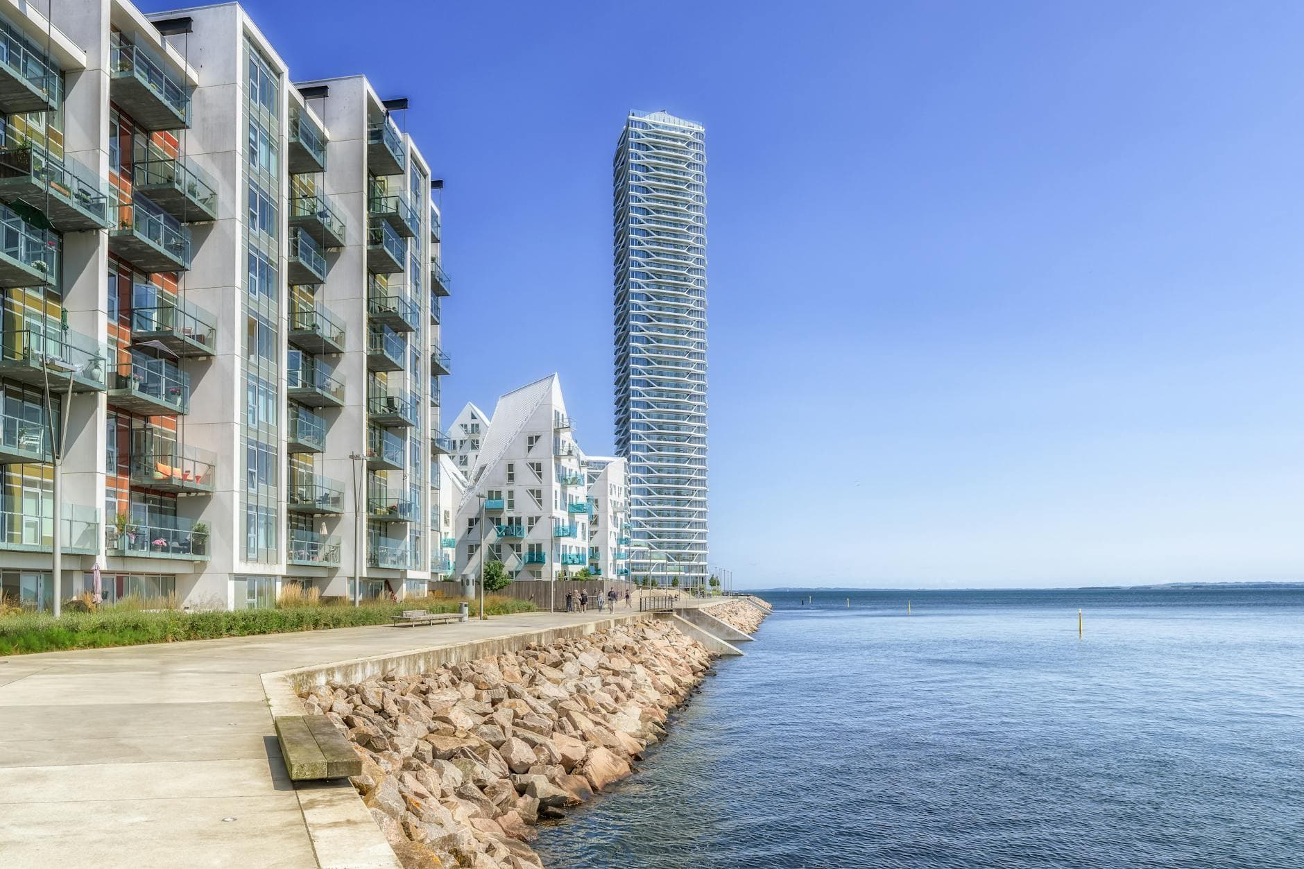 Moderne lejlighedsbyggeri og et højhus ved en havnepromenade i klart solskin.