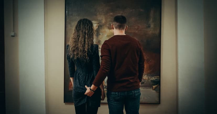 Et par set bagfra står hånd i hånd og betragter et stort maleri på en museumsvæg.