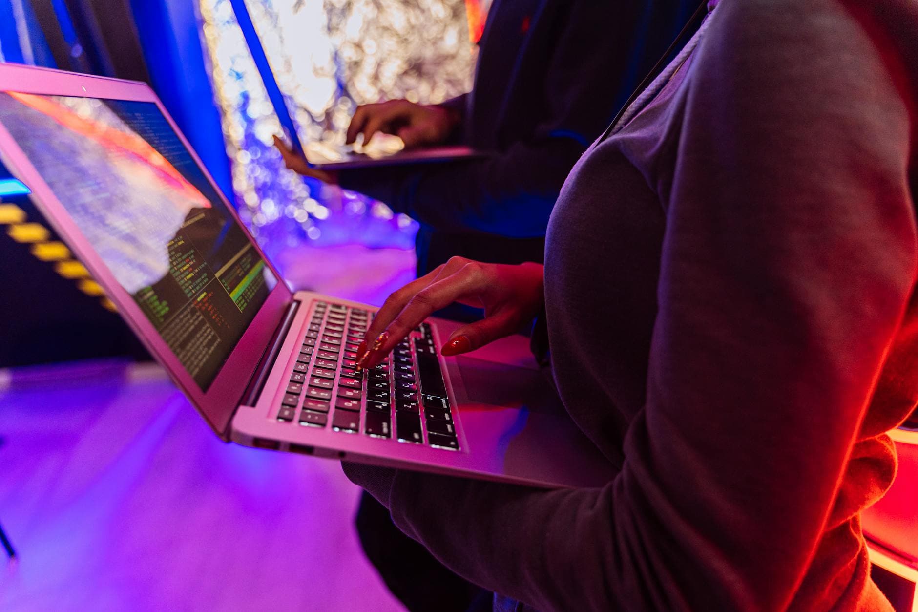 Hender skriver på en laptop med kode på skjermen i rosa og lilla neonlys.