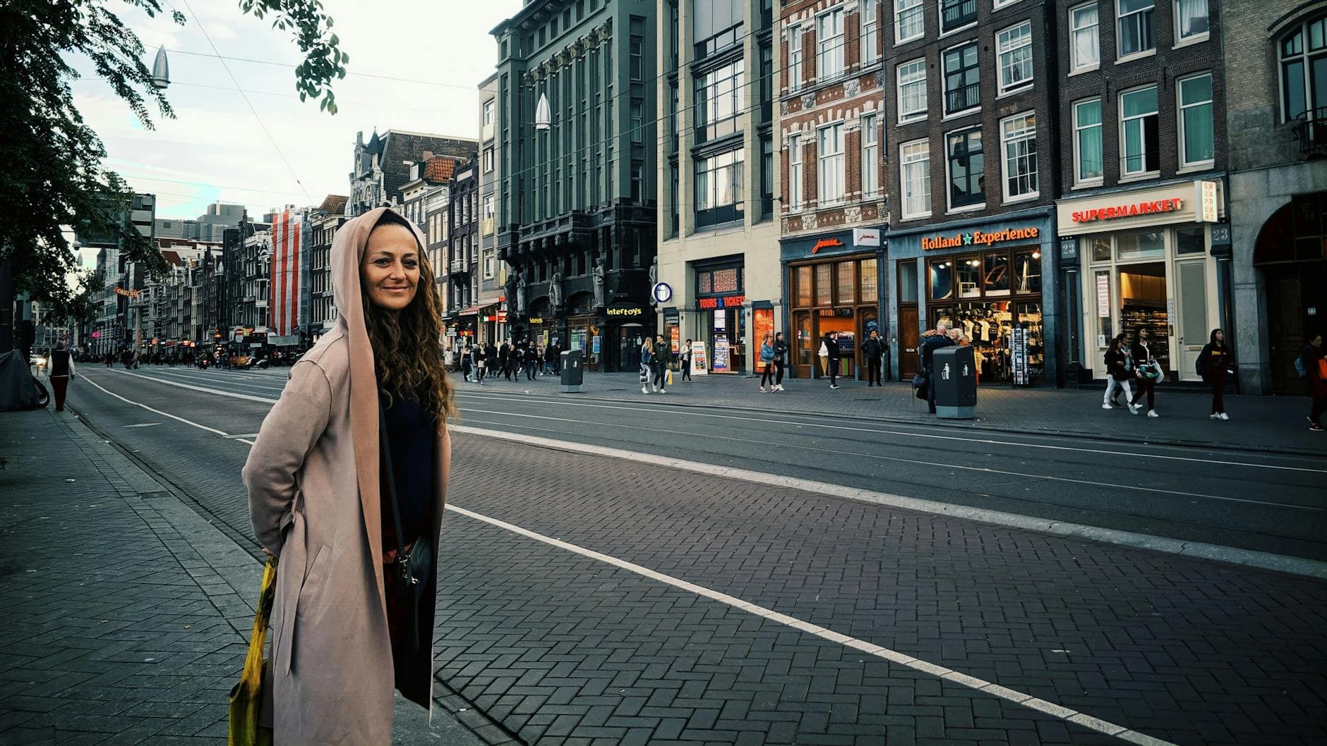 Vrouw met krullend haar in een beige jas met capuchon lacht op een brede stadsstraat.