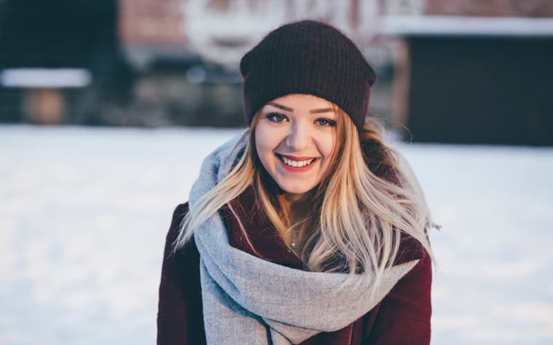 Leende person med blont hår, mörk mössa, grå halsduk och vinröd jacka i snölandskap.