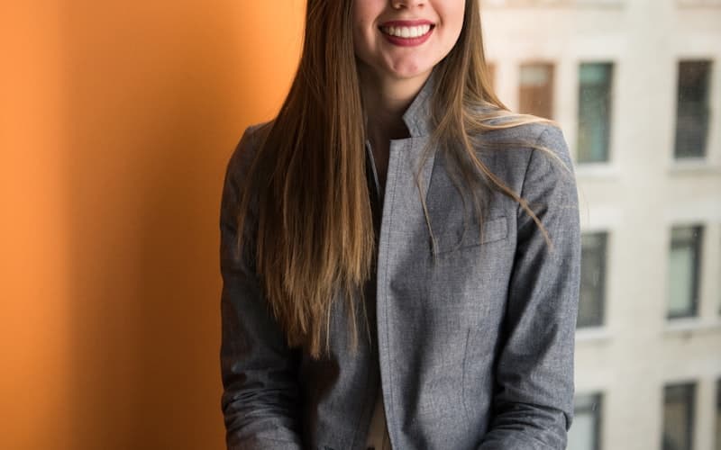 A smiling woman with long fair hair in a grey blazer stands by an orange wall.