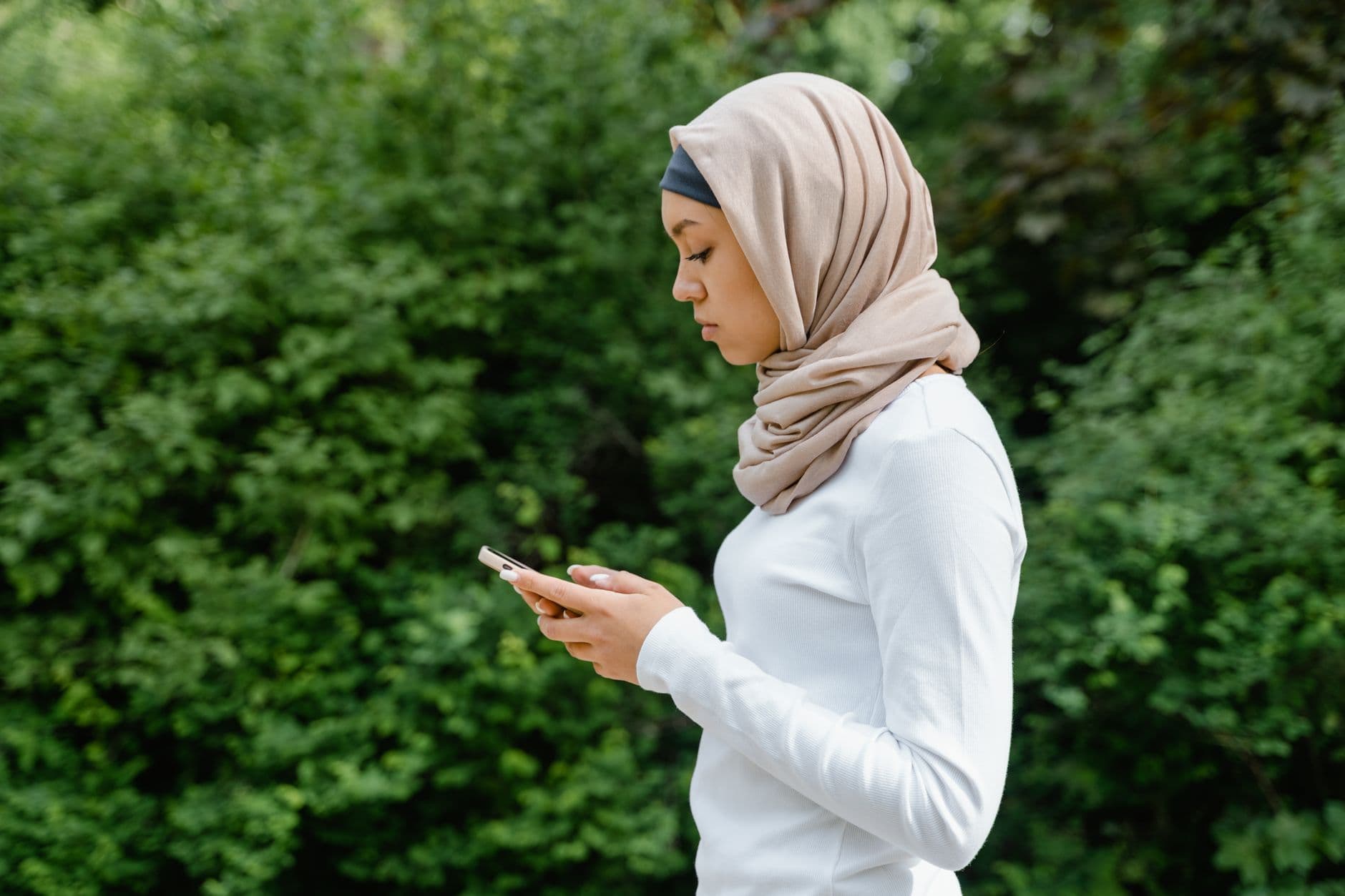 Een vrouw met een lichte hoofddoek gebruikt haar telefoon buiten, voor een groene achtergrond.