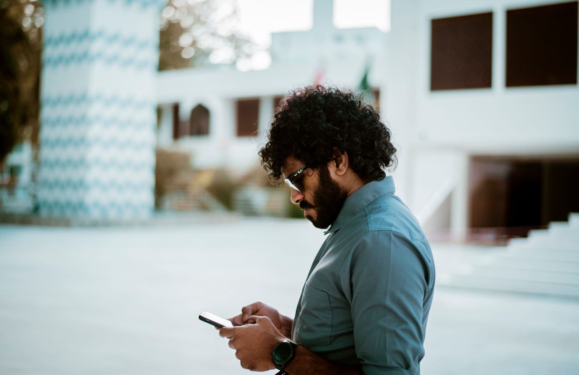 Un homme aux cheveux bouclés et à la barbe, portant des lunettes, consulte son téléphone dehors.